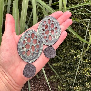 May include: A pair of oval earrings with a distinctive design. The earrings have a grey and green marbled pattern with cut-out holes. Each earring has a textured, dark grey, teardrop-shaped dangle. The earrings are displayed against a natural background.