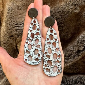 May include: A pair of dangle earrings with a black circular stud and a white and black patterned teardrop shape. The earrings have a unique design with multiple oval cutouts. The earrings are shown against a brown background.