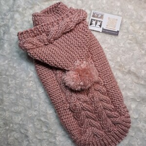 May include: A pink knitted dog sweater with a hood and a pom-pom. The sweater has a cable knit pattern.