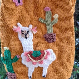 May include: Mustard yellow knitted pet sweater with a llama and cactus design. The sweater features a white llama with a colorful blanket, pink accents, and two green cacti. A pink butterfly is also included.