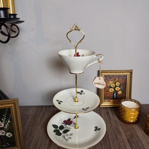 Könnte beinhalten: Eine zweistöckige Etagere mit einer weißen Porzellantasse auf der oberen Ebene und einer passenden weißen Porzellanuntertasse auf der unteren Ebene. Beide Teile sind mit dunkelroten Rosenmotiven und grünen Blättern verziert und durch einen goldfarbenen Metallgriff verbunden.