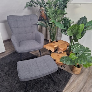 Könnte beinhalten: Ein grauer, gepolsterter Sessel und ein passender Hocker mit schwarzen Metallbeinen stehen auf einem dunkelgrauen Teppich. Ein kleiner Beistelltisch aus Holz und Topfpflanzen sorgen für einen Hauch von Natur. Der Stuhl hat eine hohe Rückenlehne.