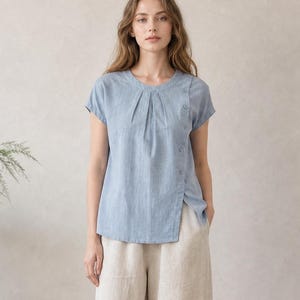 May include: A light blue short-sleeved blouse with a round neckline, vertical pleats at the chest, and a side button closure, paired with wide-leg beige trousers. The blouse features a subtle textured fabric and a slightly asymmetrical hemline.