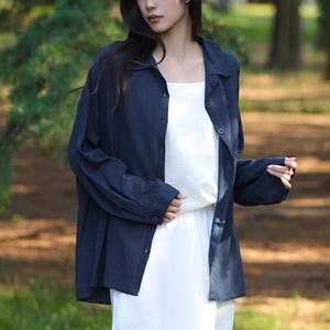 May include: A navy blue button-down shirt worn over a white dress. The shirt has long sleeves and a relaxed fit. The background is a blurred outdoor scene with trees and natural light. The overall style is casual and comfortable.
