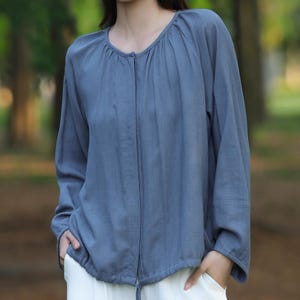 May include: A long-sleeved, slate blue blouse with a gathered neckline and a drawstring waist is paired with white wide-leg pants. The outfit is made of a soft, flowing fabric, perfect for a casual, comfortable look. The model is standing outdoors in a park setting.