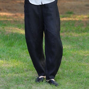 May include: Black linen pants with a relaxed fit. The pants have a wide leg and are worn with a light-colored button-down shirt. The person is standing on green grass.