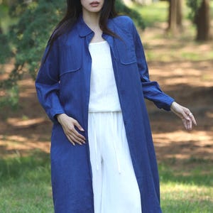 May include: A long, dark blue linen shirt-dress with two front pockets, worn over a white top and wide-leg trousers. The outfit is buttoned down the front and has long sleeves. The model is standing in a grassy outdoor setting.