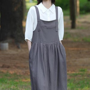 May include: A woman wearing a white collared shirt under a gray overall dress. The dress has a gathered skirt and pockets. The outfit is made of a soft, flowing fabric. The background is a blurred view of a forest with trees.