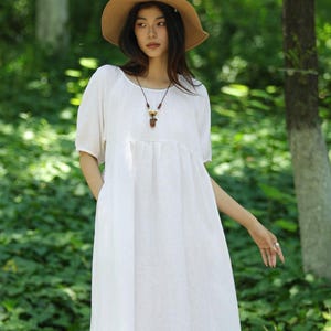Puede incluir: Una mujer con un vestido de lino blanco de manga corta y cuello redondo. Lleva un sombrero de paja y un collar con colgante. El vestido llega a media pantorrilla. El fondo es un bosque verde.
