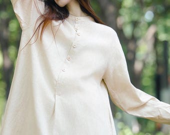以下が含まれることがあります： ベージュのリネンドレス。マンダリンカラーとボタンディテールが特徴。長袖でゆったりとしたフィット感。モデルは屋外で着用。ドレスは足首まで届きます。