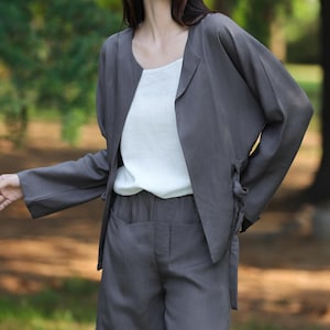 May include: A charcoal gray linen jacket and shorts set. The jacket has long sleeves and a tie closure. The shorts have pockets. A white top is worn underneath the jacket. The outfit is worn outdoors in a park setting.
