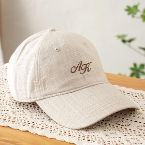 Op de afbeelding: Beige baseballpet met de geborduurde letters "AK" in bruin script. De pet is gemaakt van een textuurstof en ligt op een decoratieve kanten loper op een houten oppervlak. De achtergrond is zacht vervaagd.