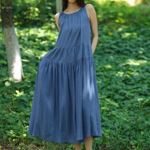 Può includere: Un abito midi blu senza maniche con gonna a balze e scollo tondo. L'abito arriva a metà polpaccio. La modella indossa sandali bianchi e si trova in un ambiente naturale e verde. L'abito è realizzato in un tessuto morbido e fluido.