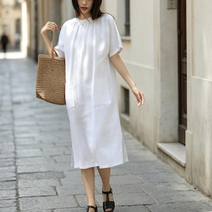May include: A woman in a white linen dress with short sleeves and two front pockets, carrying a woven tote bag. The dress reaches mid-calf, paired with black sandals. The setting is a stone-paved street, with buildings on either side.