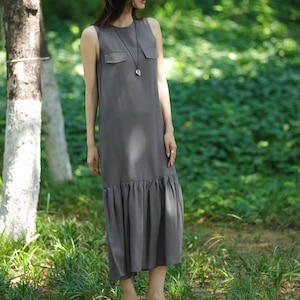May include: A sleeveless, gray midi dress with a round neckline and two small chest pockets. The dress features a ruffled hem and is paired with black flats and a silver pendant necklace. The model is standing outdoors in a green, natural setting.