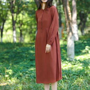 May include: A long-sleeved, rust-colored dress with a button-down front and a modest, flowing silhouette. The dress features a round neckline and is made of a soft, lightweight fabric. The model is standing in a natural outdoor setting with trees and greenery.