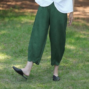 May include: Green linen pants with a scalloped hem. The pants are a solid, deep green color and appear to be made of a lightweight fabric. The pants are cropped and worn with black flats. The background is a grassy area.
