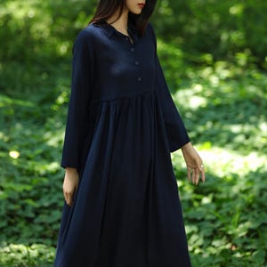 Peut inclure: Robe bleu marine à manches longues avec col et boutonnage sur le devant. La robe a une taille froncée et une jupe ample. Le modèle se trouve dans un cadre forestier avec un feuillage vert en arrière-plan.