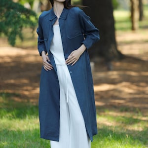 May include: A woman wearing a long, dark blue linen coat over a white top and pants. The coat has a collared neckline and button closures. The outfit is worn outdoors in a park setting with green grass and trees in the background.