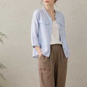 May include: A light blue button-down shirt with a collarless neckline and two chest pockets, worn over a plain white tank top. The outfit includes brown cropped trousers with a relaxed fit and beige slip-on shoes with a woven texture.
