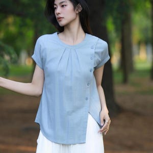 May include: Light blue short-sleeve top with a round neckline and a pleated front. The top features a buttoned asymmetrical closure on the side. The garment is paired with white pants, creating a casual and stylish outfit.