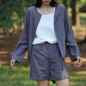May include: A three-piece outfit in a natural setting. The ensemble includes a gray jacket with long sleeves, a white top, and gray shorts. The jacket has a unique collarless design and the shorts have pockets.