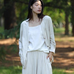 Puede incluir: Conjunto de lino beige claro compuesto por una blazer, un top y un pantalón de pernera ancha. La blazer tiene dos botones y bolsillos. El top es de estilo cuello cuadrado. El pantalón tiene un diseño plisado. El conjunto se lleva al aire libre.