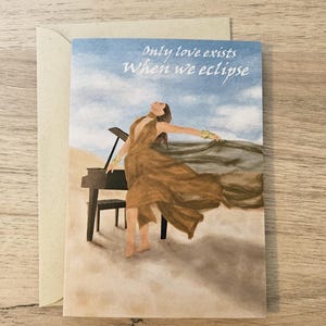 Op de afbeelding: Wenskaart met een aquarel illustratie van een persoon in een vloeiende bruine jurk bij een piano. De kaart heeft de tekst "Only love exists When we eclipse" op een blauwe hemel achtergrond. De kaart zit in een beige envelop.