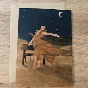 Op de afbeelding: Een wenskaart met een aquarel schilderij van een persoon in een vloeiende bruine jurk, met uitgestrekte armen, bij een piano onder een sterrenhemel met een halve maan. De kaart zit in een beige envelop.