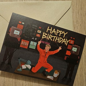 May include: A birthday card with a retro-style illustration. The card features a person in an orange jumpsuit celebrating in front of a control panel. The words "HAPPY BIRTHDAY" are written in yellow. A brown envelope is behind the card.