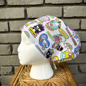 Pode incluir: Touca cirúrgica branca com patches de personagens de desenhos animados coloridos. Os patches incluem personagens como Badtz-Maru, Pekkle e Chococat, juntamente com arco-íris, sorvetes e notas musicais. A touca é amarrada na parte de trás.