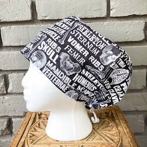 Puede incluir: Un gorro de quirófano negro y blanco con un diseño de calavera y huesos cruzados. El gorro presenta términos anatómicos como "cráneo", "costillas", "fémur", "húmero" y "vértebras".