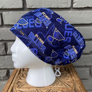 May include: A navy blue scrub cap featuring the St. Louis Blues logo and team name in white and light blue. The cap is patterned with the team's logo and name, and has a white drawstring for adjustment. The cap is displayed on a white mannequin head.