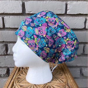 Puede incluir: Un gorro de quirófano azul con un patrón de calaveras de azúcar de colores. El gorro tiene un lazo en la parte posterior.
