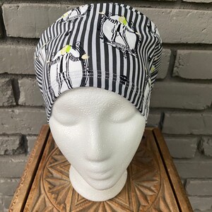 May include: Black and white striped scrub hat with a repeated pattern of a cartoon character wearing a black and white striped suit. The character is holding a scythe and has a green face.