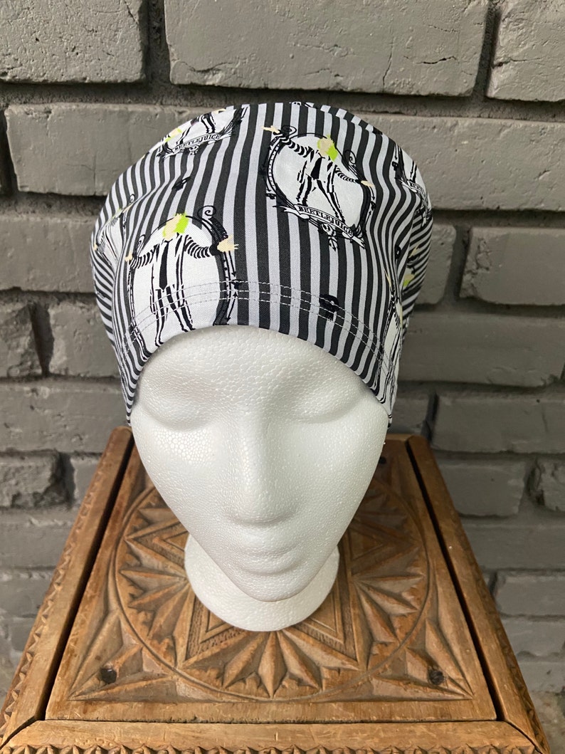 May include: Black and white striped scrub hat with a repeated pattern of a cartoon character wearing a black and white striped suit. The character is holding a scythe and has a green face.