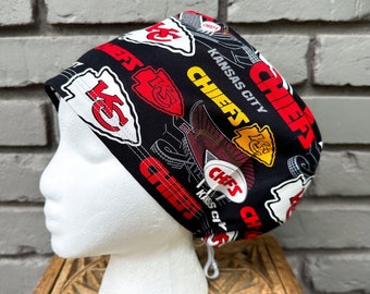 May include: A black scrub cap featuring the Kansas City Chiefs logo in red, white, and yellow. The cap has a drawstring and is displayed on a white mannequin head. The words "CHIEFS" and "KANSAS CITY" are also visible.