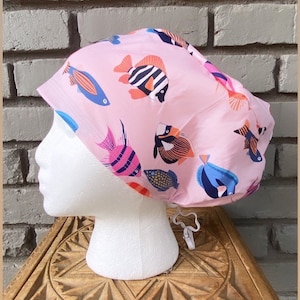 May include: A pink surgical scrub hat with a colourful fish print. The hat has a drawstring tie.