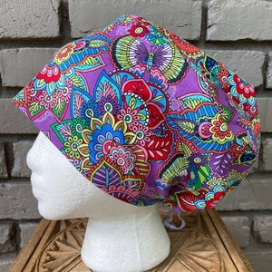 May include: A colorful scrub cap featuring a vibrant floral and paisley pattern in shades of purple, blue, red, green, and yellow. The cap is designed to fit comfortably and has adjustable ties for a secure fit. The cap is displayed on a white mannequin head.