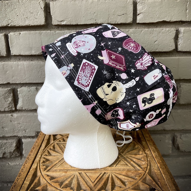 May include: A black and white patterned surgical scrub hat with a skull, stars, and the words "Magic Spells".