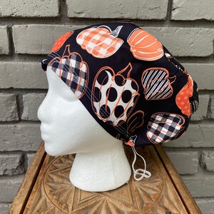 May include: A black scrub hat with a tie string. The hat has a pattern of orange pumpkins with black and white polka dots, gingham, and stripes.