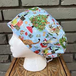May include: A light blue surgical scrub cap featuring a festive Christmas cat design. The cap is patterned with Christmas trees, snowflakes, and various cats wearing Santa hats. The cap is made of a soft, breathable fabric.