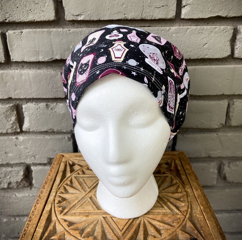 May include: Black scrub hat with a pink and white print of various potion bottles, tarot cards, and a black cat. The print is on a black background with white stars.
