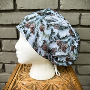 May include: A patterned scrub cap featuring a winter scene with pine cones and snow-covered branches. The cap is primarily white and blue with brown pine cones. The cap is displayed on a white mannequin head.