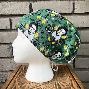 May include: Green surgical scrub hat with a Christmas-themed print featuring a cartoon character with the text "Cotton Headed Ninny" repeated throughout the design.