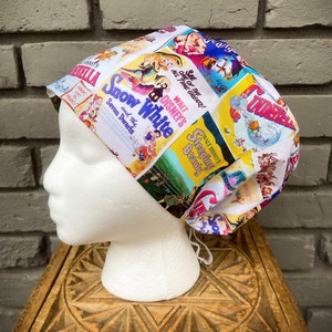 以下が含まれることがあります： 白地に、白雪姫と7人の小人、眠れる森の美女、シンデレラなど、ヴィンテージのディズニー映画のポスターがカラフルにプリントされたスクラブハット。ポスターは鮮やかな色使いで、太字のテキストが特徴です。