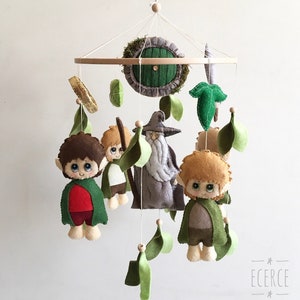 Puede incluir: Móvil infantil caprichoso con personajes de fieltro y elementos de una historia de fantasía. El móvil incluye figuras de hobbits, un mago y una puerta, con hojas verdes y detalles de madera. Los personajes visten de verde, rojo y marrón.