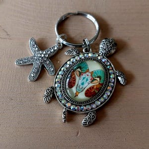 Könnte beinhalten: Ein silberfarbener Schlüsselanhänger mit einem Schildkrötenanhänger mit farbenfrohem Design und einem Seestern-Charm. Der Schildkrötenanhänger hat einen dekorativen Rand aus schillernden Strasssteinen. Der Schlüsselanhänger liegt auf einer Holzoberfläche.