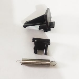 Könnte beinhalten: Drei schwarze Kunststoffteile für einen Türschlossmechanismus. Die Teile umfassen eine Feder, ein kleines rechteckiges Stück mit zwei Löchern und ein größeres Stück mit einem gebogenen Arm.