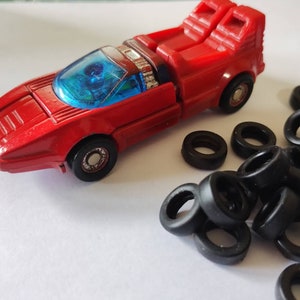 Puede incluir: Un coche de juguete rojo con ruedas negras y un interior azul. El coche está rodeado de una colección de anillos de goma negra.
